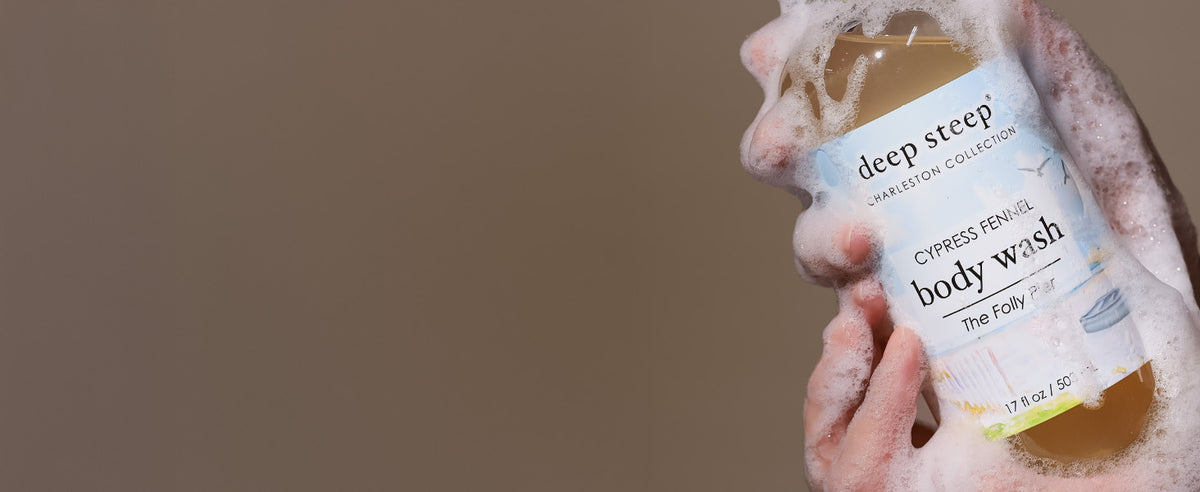 A hand wrapped in white foam holds a bottle of Deep Steep Cypress Fenel Body Wash against a plain taupe background.