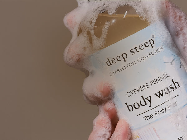 A close-up of a hand holding a bottle of Deep Steep Cypress Fennel Body Wash, covered in thick white foam, against a solid taupe background.