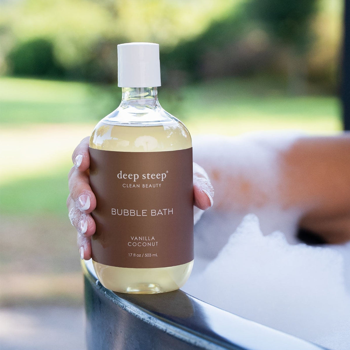 A hand with white nail polish holds a bottle of deep steep Clean Beauty Bubble Bath in Vanilla Coconut. The clear bottle shows a pale yellow liquid inside and has a brown label with a white cap.
