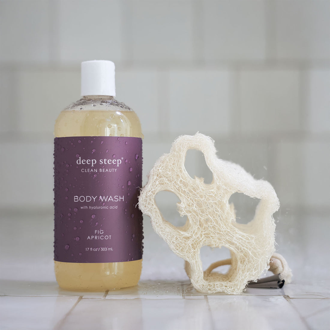 Bottle of Deep Steep Fig Apricot Body Wash with a loofah on a tiled bathroom floor
