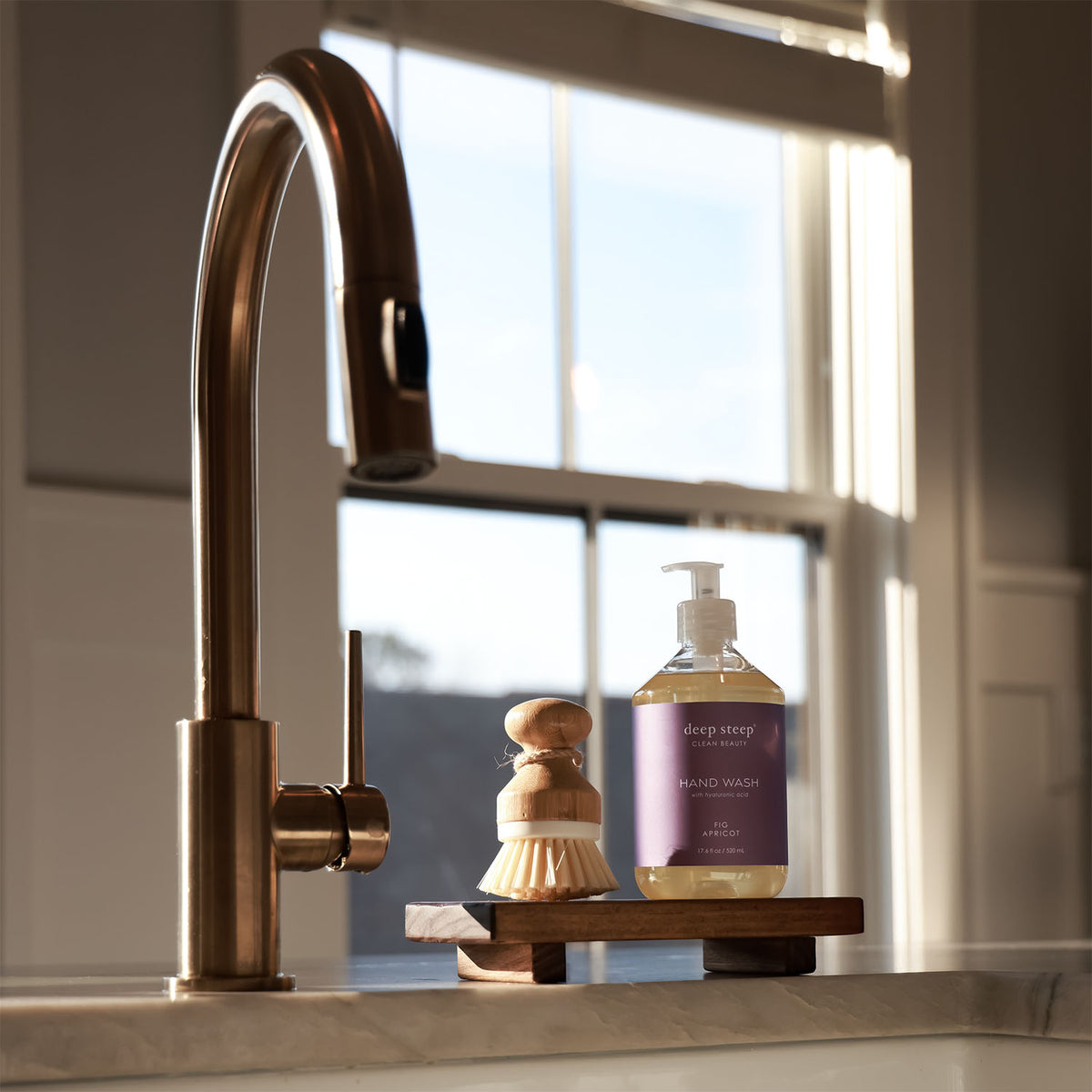 Kitchen sink area with a gold faucet, wooden brush, and a bottle of Deep Steep Fig Apricot Liquid Hand Wash on a wooden stand