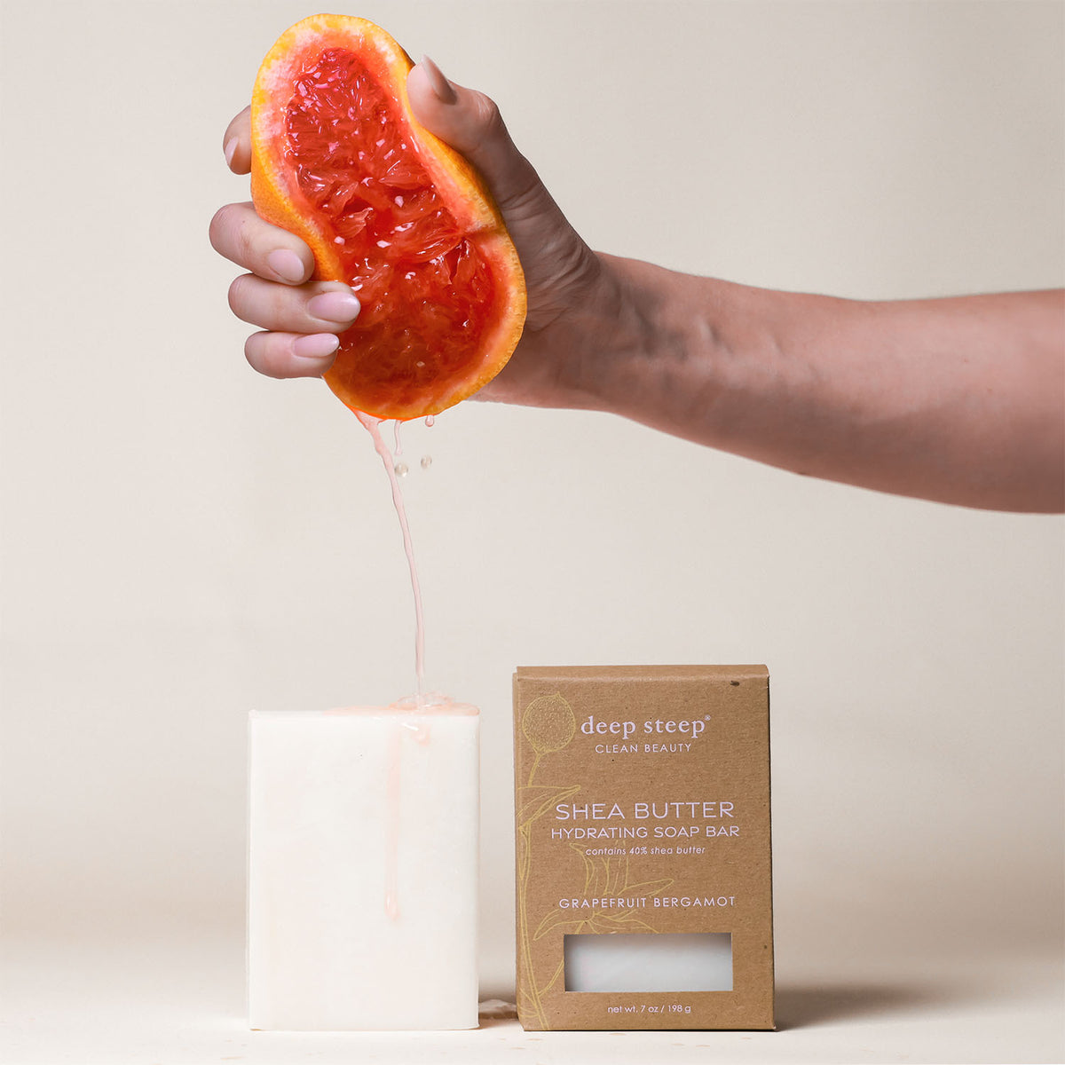 7 oz Deep Steep Grapefruit Bergamot hydrating bar soap containing 40% shea butter alongside a hand squeezing a fresh grapefruit half on a neutral background