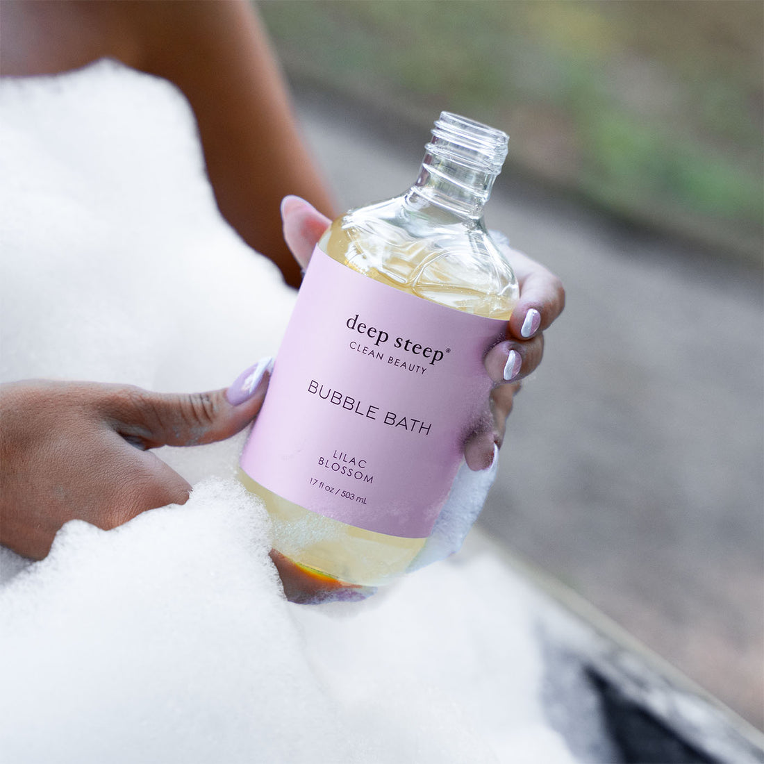 17 oz / 503 ml Deep Steep Lilac Blossom bubble bath in a bottle with a light purple label being held by someone surrounded by bubbles