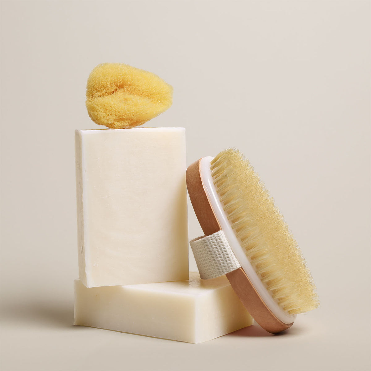 7 oz Deep Steep hydrating bar soap containing 40% shea butter stacked with a bath sponge and a wooden body brush on a neutral background
