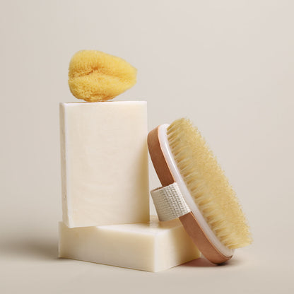 7 oz Deep Steep hydrating bar soap containing 40% shea butter stacked with a bath sponge and a wooden body brush on a neutral background