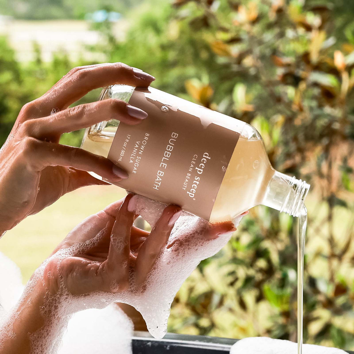 17 oz / 503 ml Deep Steep Brown Sugar Vanilla bubble bath being poured from a bottle into soapy hands against a natural outdoor background