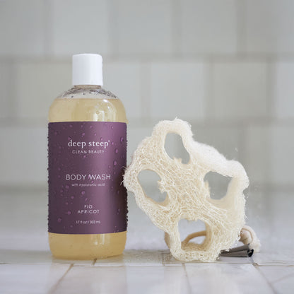 Bottle of Deep Steep Fig Apricot Body Wash with a loofah on a tiled bathroom floor
