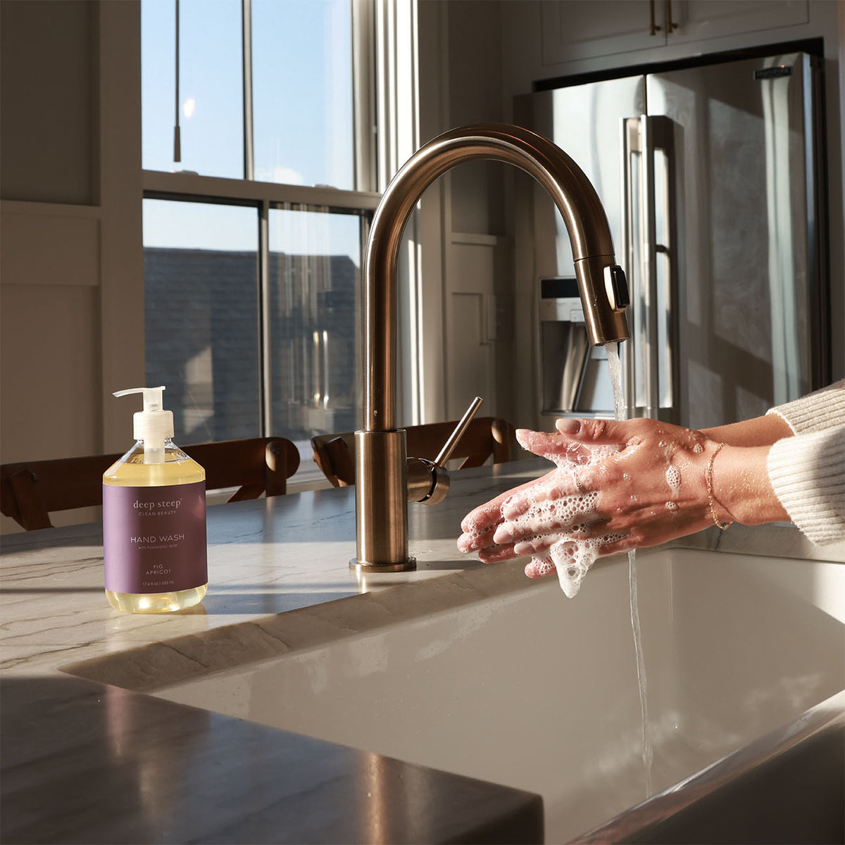 Person washing hands with soap under a running faucet, next to a bottle of Deep Steep Fig Apricot Liquid Hand Wash, 17.6 fl oz bottle on a countertop