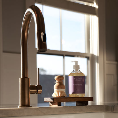 Kitchen sink area with a gold faucet, wooden brush, and a bottle of Deep Steep Fig Apricot Liquid Hand Wash on a wooden stand