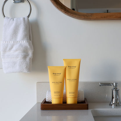 Two tubes of Deep Steep Grapefruit Bergamot body lotion and body butter on a wooden tray on a bathroom counter