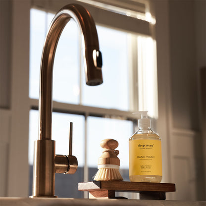 Kitchen sink area with a gold faucet, wooden brush, and a bottle of Deep Steep Grapefruit Bergamot Liquid Hand Wash on a wooden stand