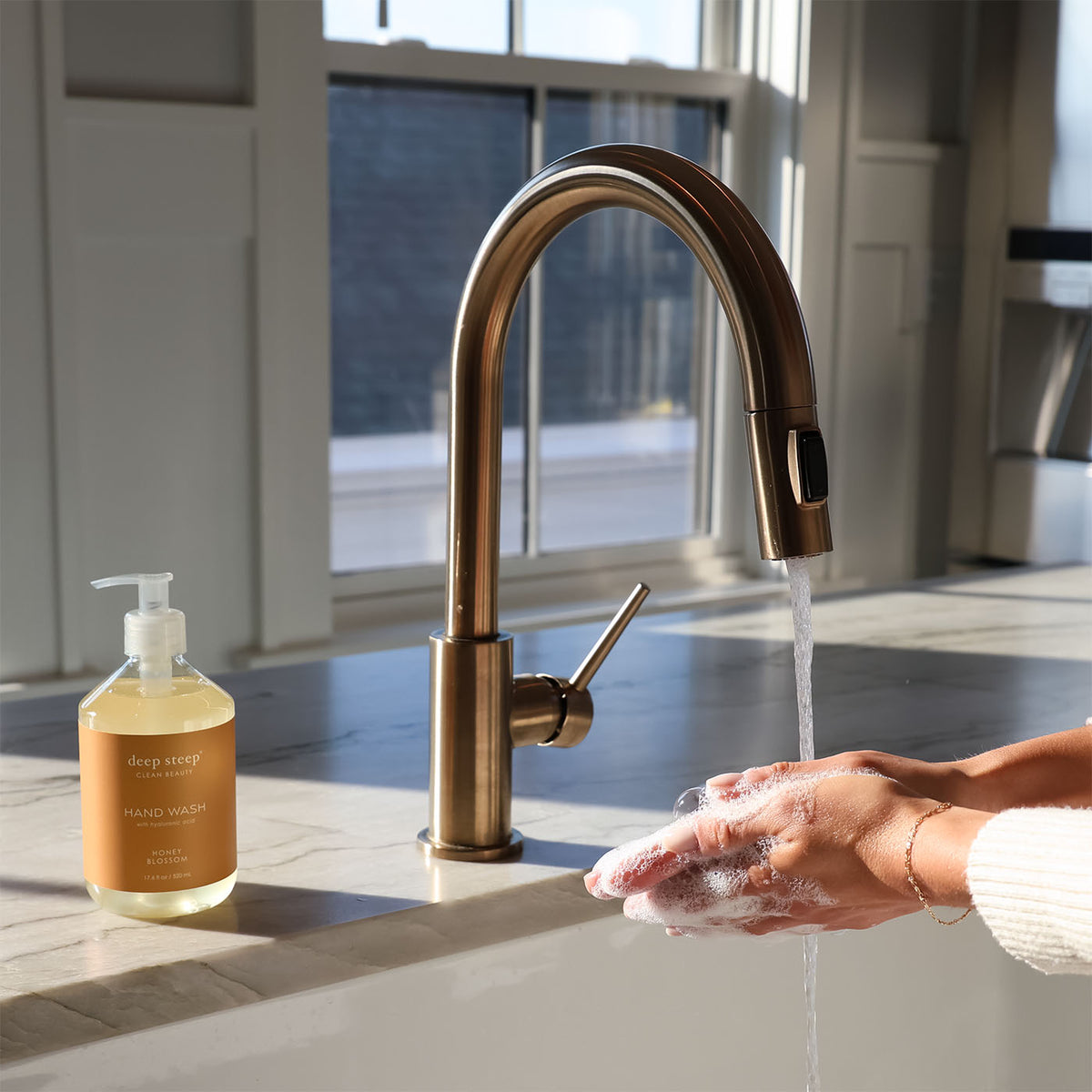 Person washing hands with soap under a running faucet, next to a bottle of Deep Steep Honey Blossom Liquid Hand Wash, 17.6 fl oz bottle on a countertop