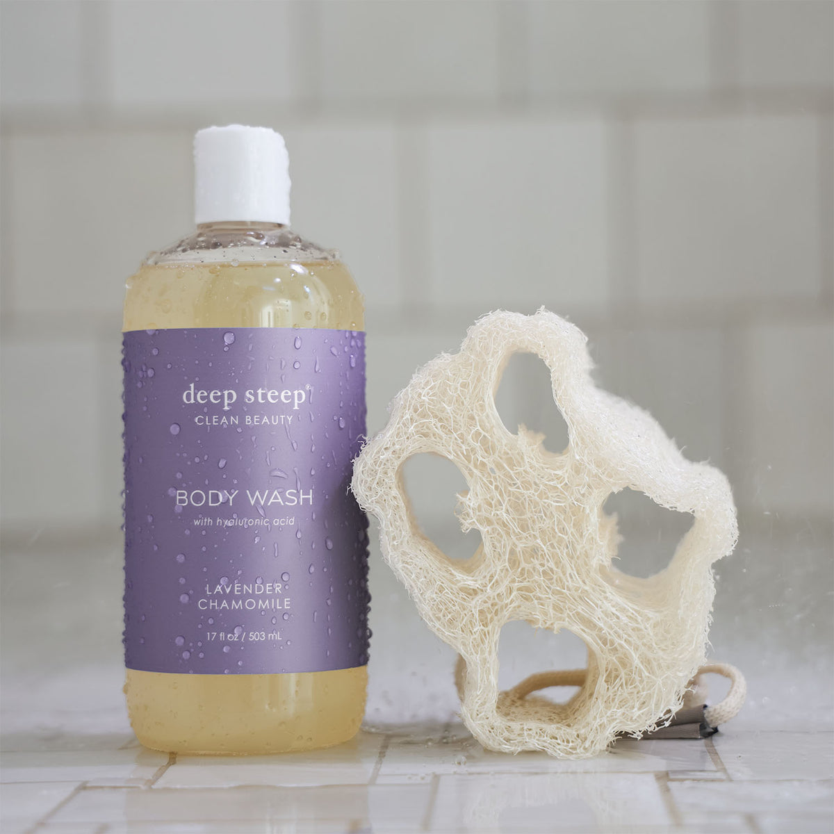 Bottle of Deep Steep Lavender Chamomile Body Wash with a loofah on a tiled bathroom floor