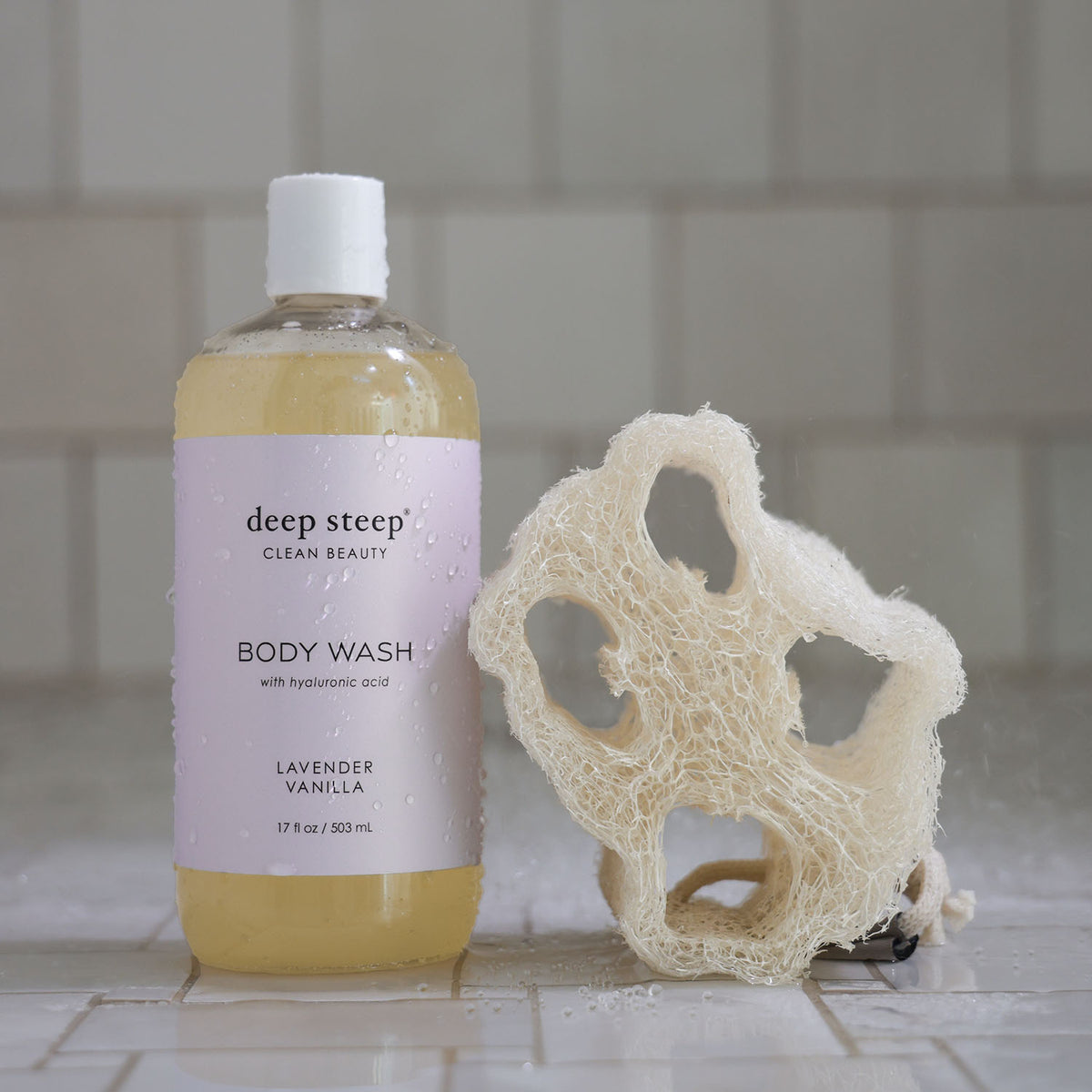 Bottle of Deep Steep Lavender Vanilla Body Wash with a loofah on a tiled bathroom floor