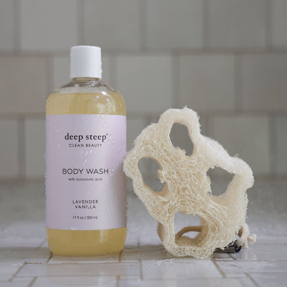 Bottle of Deep Steep Lavender Vanilla Body Wash with a loofah on a tiled bathroom floor
