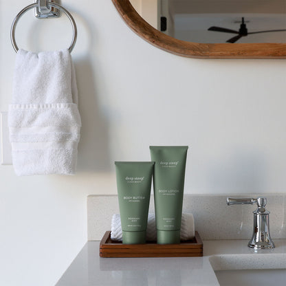 Two tubes of Deep Steep Rosemary Mint body lotion and body butter on a wooden tray on a bathroom counter