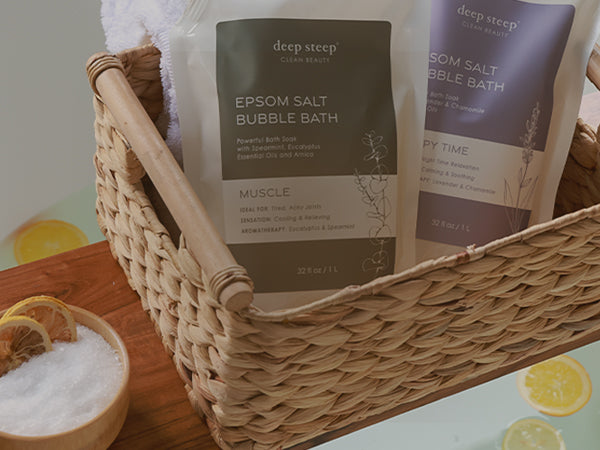 Two Deep Steep Epsom Salt Bubble Bath bags displayed in a woven wicker basket, with a small bowl of bath salt and lemon slices nearby on a wooden surface.